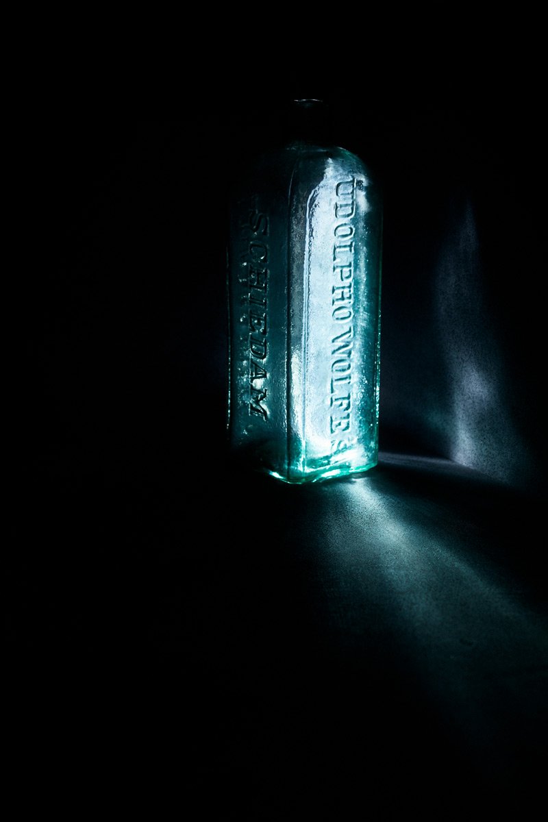 An embossed aqua bottle lit from within, casting light across the surface, Glass by Hannah Caldwell