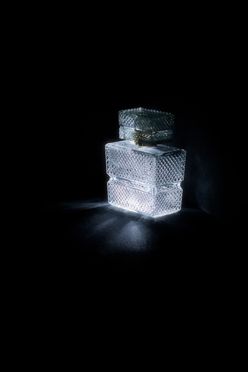 A square faceted decanter and stopper glowing white against black, Glass by Hannah Caldwell