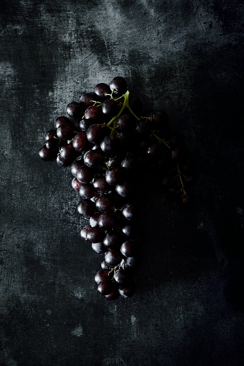 A bunch of dark grapes against black, RAW by Hannah Caldwell