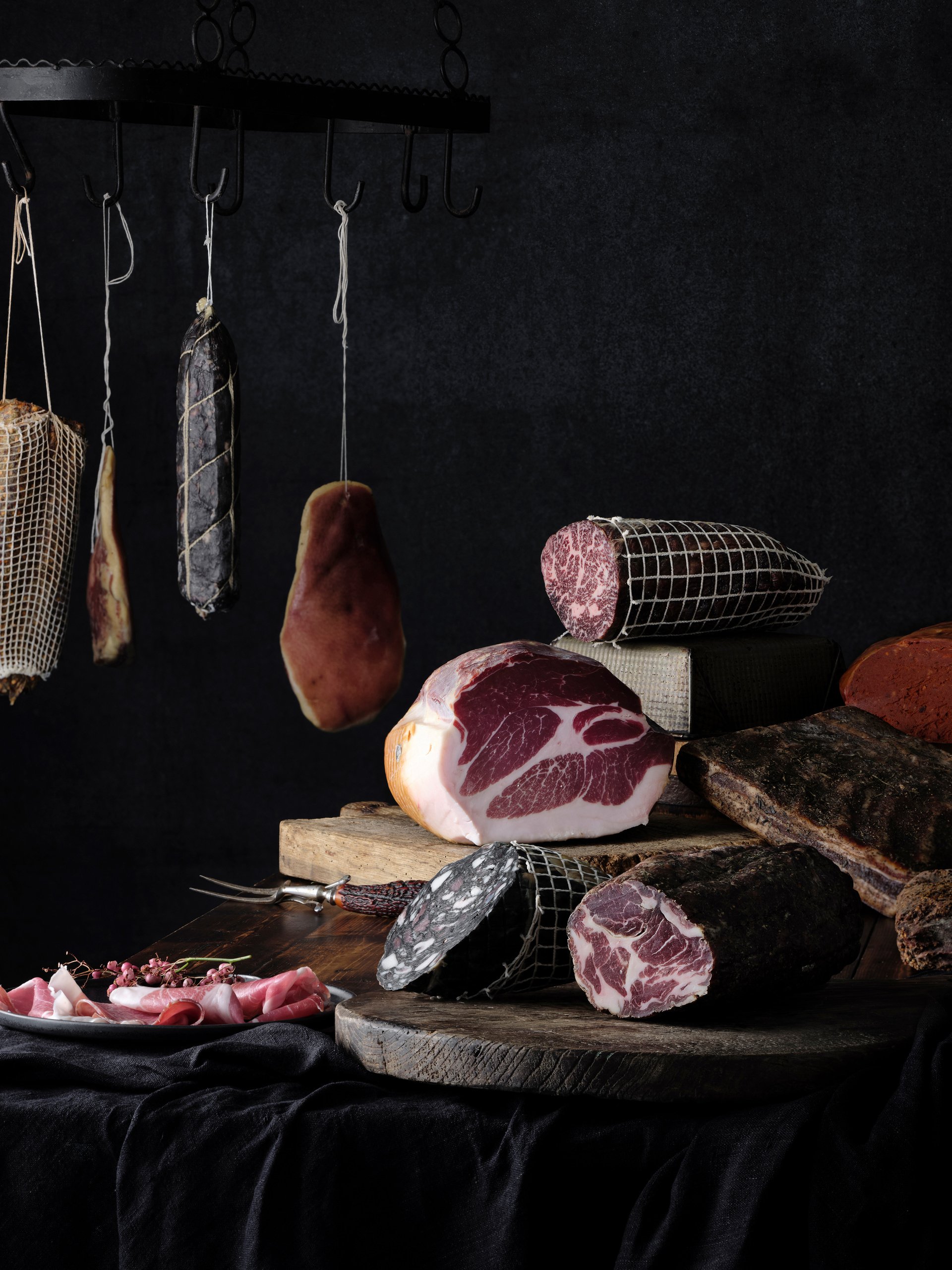 Hanging cured meats and salumi on a dark wall, a whole coppa and sliced meats on boards below, Savour and Grace by Hannah Caldwell