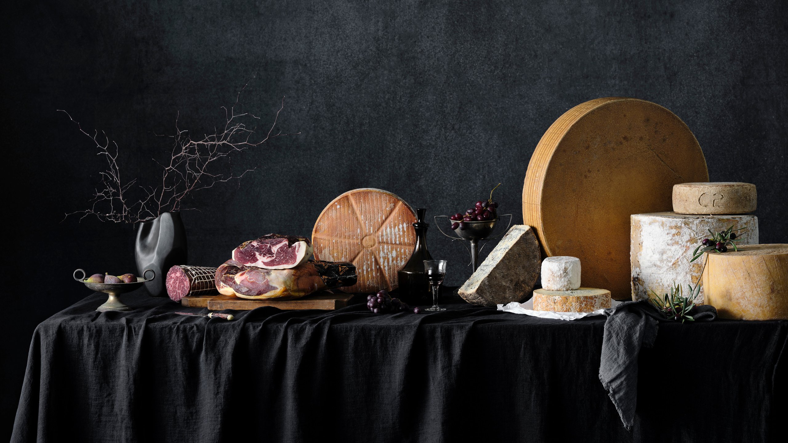 A draped black table set with prosciutto, a wheel of cheese, small dark grapes and a glass, Savour and Grace by Hannah Caldwell
