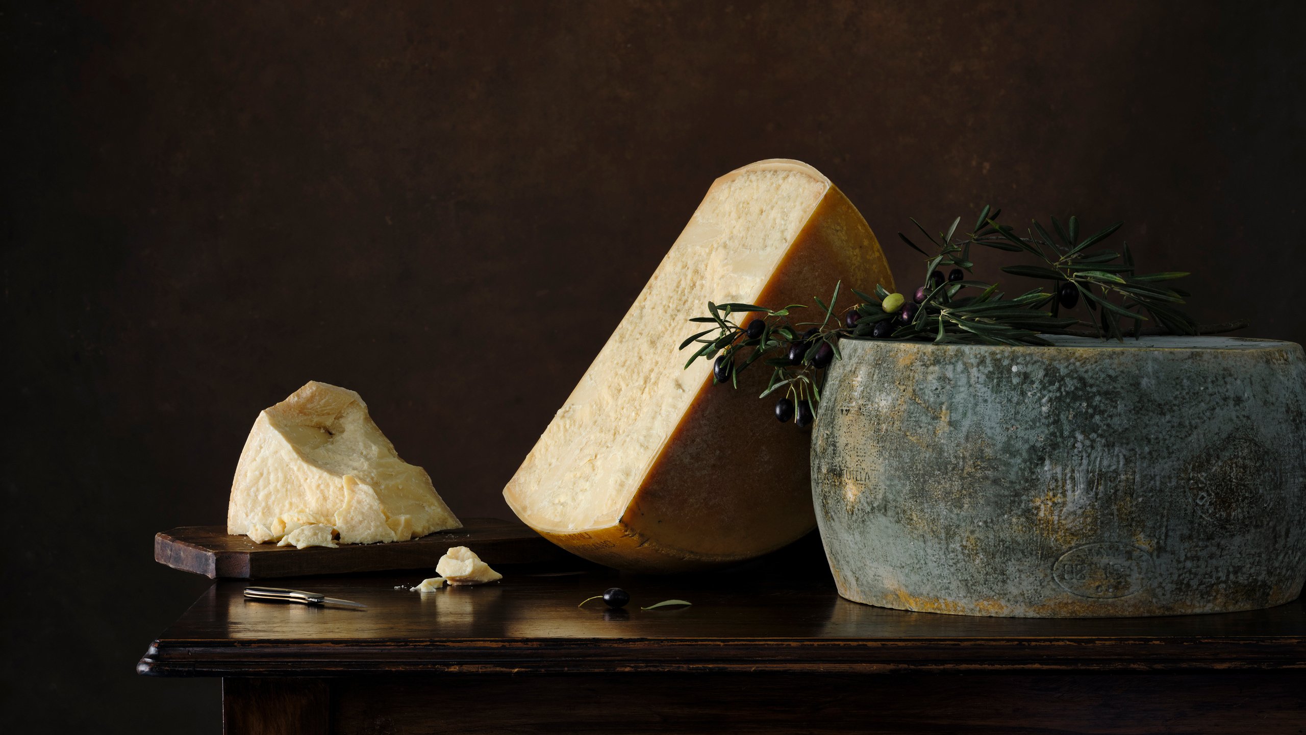 A large wedge of aged parmesan leaning against a mossy green pot with olive branches, a glass of red wine alongside, Savour and Grace by Hannah Caldwell